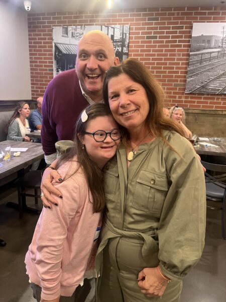 A young woman with Down Syndrome, Cara Cushing, in a pink jacket with glasses with another woman and man all smiling at a networking event.